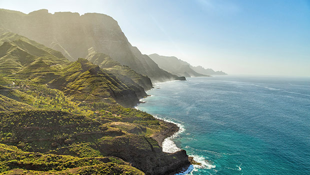 Dramatiska kustklippor täckta av grönskande vegetation möter det azurblå havet under en klar himmel i mjukt solljus.