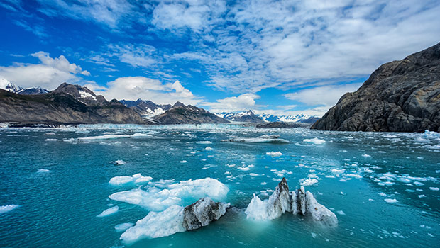 Isflak flyter på ett turkost glaciärhav, omgivet av snötäckta berg och dramatiska klippor under en delvis molnig himmel.