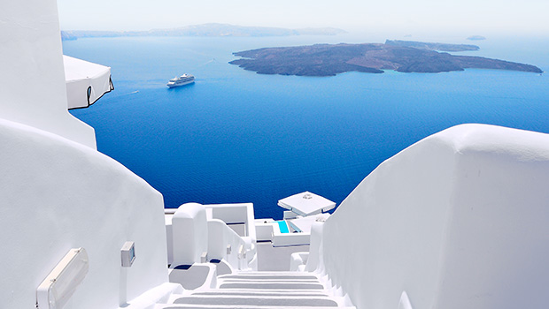 Vitkalkade trappor leder ner mot det djupblå havet i Santorini, med ett kryssningsfartyg och vulkaniska öar i bakgrunden.