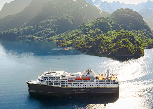 Ett Havila Kystruten-fartyg seglar genom en naturskön fjord, omgivet av grönskande berg och spegelblankt vatten.