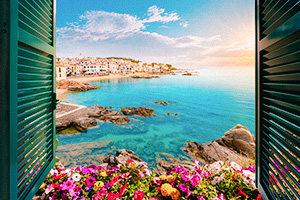 Utsikt mot havet och en kustby i Spanien genom ett öppet fönster med blommor.
