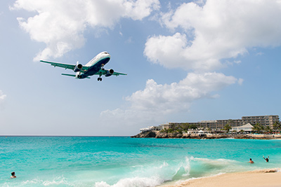 Ett flygplan flyger över en tropisk strand och hotell i bakgrunden.