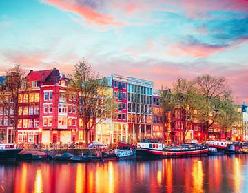 Kvällsvy över Amsterdam med kanaler, husbåtar och upplysta byggnader vid vattnet.